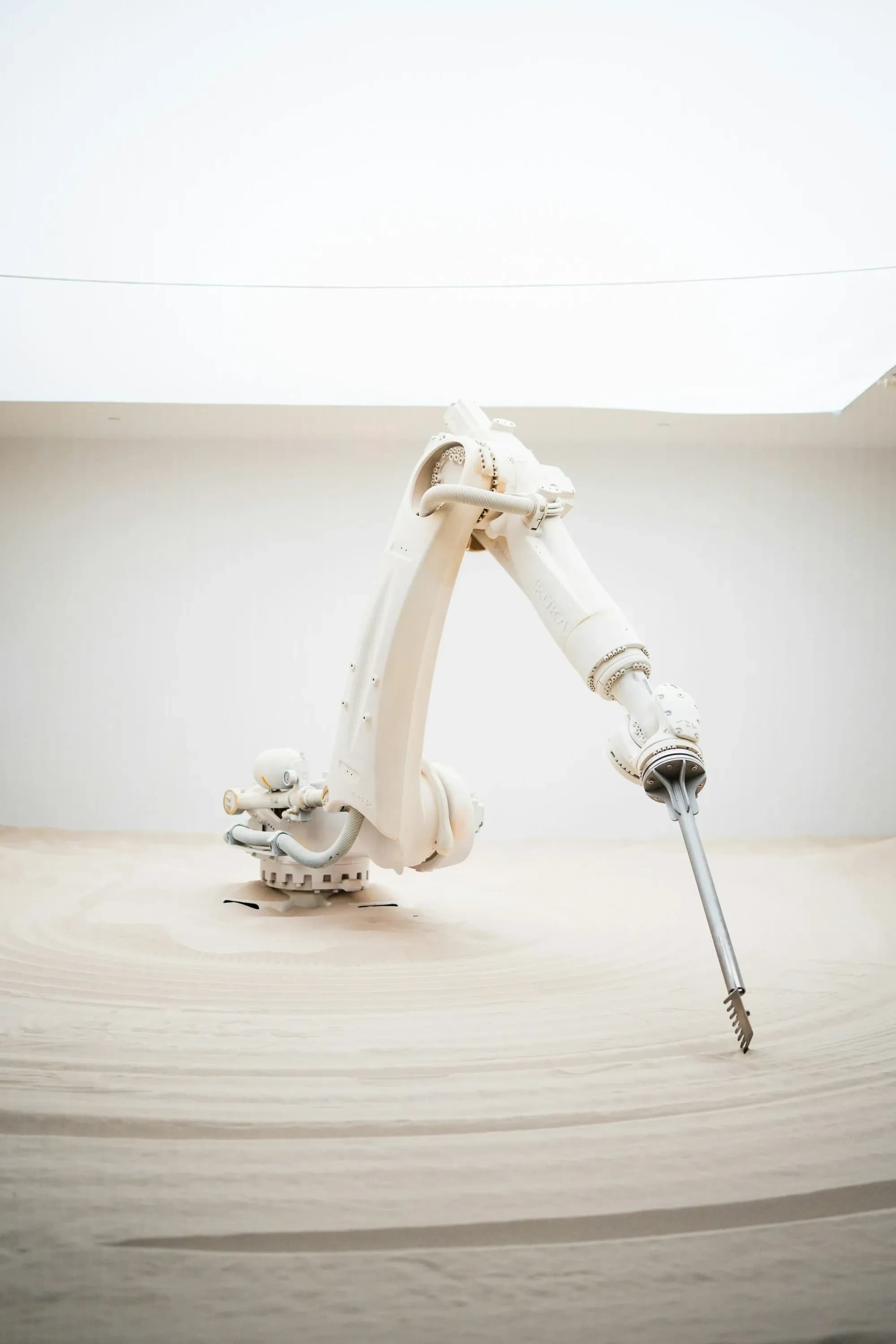 Weißer Roboterarm zieht mit einem Stab Linien in hellen Sand